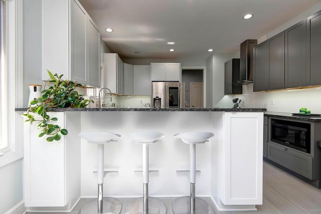 kitchen featuring dark stone counters, stainless steel fridge with ice dispenser, gray cabinetry, black microwave, and wall chimney exhaust hood