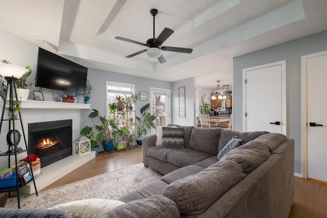 living room with ceiling fan with notable chandelier, light hardwood / wood-style floors, a fireplace, and a raised ceiling