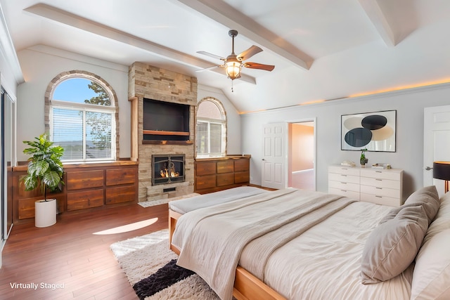 bedroom with ceiling fan, wood-type flooring, a stone fireplace, and vaulted ceiling with beams