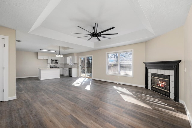 unfurnished living room featuring ceiling fan with notable chandelier, a tiled fireplace, dark wood-type flooring, and a tray ceiling