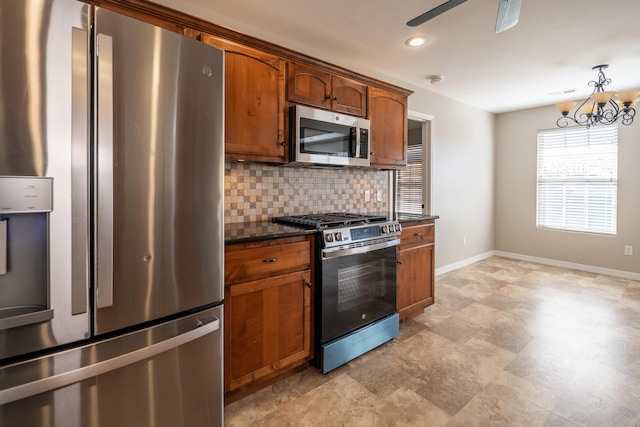 kitchen featuring dark stone countertops, pendant lighting, stainless steel appliances, ceiling fan with notable chandelier, and backsplash