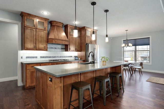 kitchen with custom exhaust hood, hanging light fixtures, a kitchen island with sink, dark wood-type flooring, and stainless steel appliances