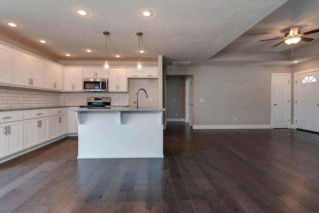 kitchen featuring white cabinetry, decorative light fixtures, appliances with stainless steel finishes, light stone countertops, and a kitchen island with sink