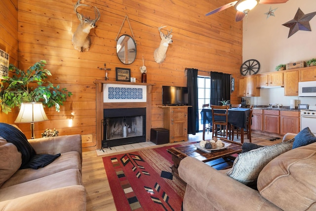 living room featuring ceiling fan, a towering ceiling, a tiled fireplace, and light wood-type flooring