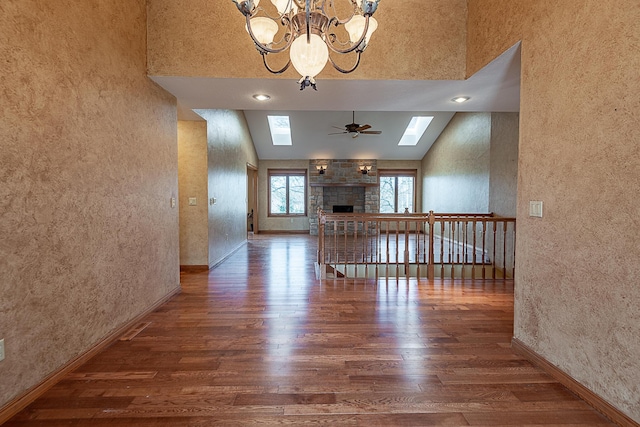 unfurnished living room with a stone fireplace, dark hardwood / wood-style floors, ceiling fan with notable chandelier, high vaulted ceiling, and a skylight