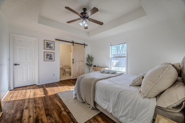 bedroom with ensuite bath, ceiling fan, wood-type flooring, a barn door, and a raised ceiling