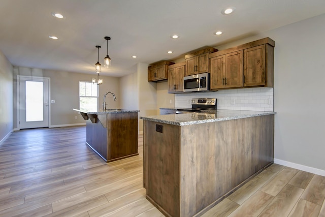 kitchen with stainless steel appliances, light stone countertops, a center island with sink, decorative light fixtures, and light wood-type flooring