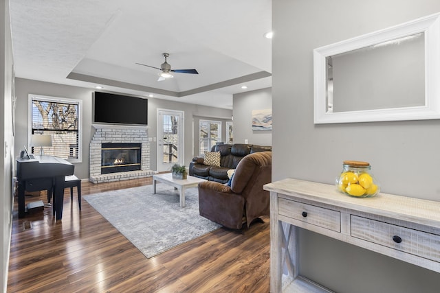 living room featuring ceiling fan, a tray ceiling, dark hardwood / wood-style flooring, and a brick fireplace