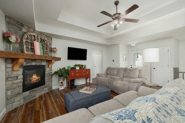 living room with ceiling fan, a tray ceiling, dark hardwood / wood-style flooring, and a stone fireplace