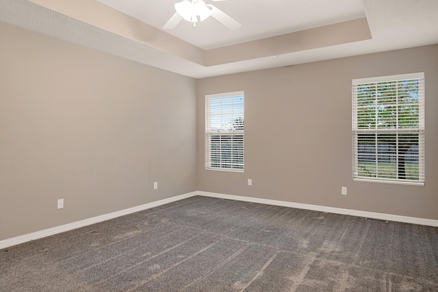 empty room with a raised ceiling, ceiling fan, a healthy amount of sunlight, and dark carpet