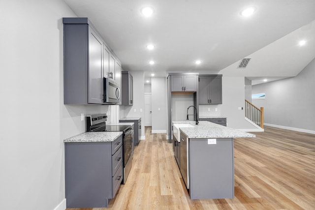 kitchen featuring gray cabinets, appliances with stainless steel finishes, a center island with sink, and light stone counters