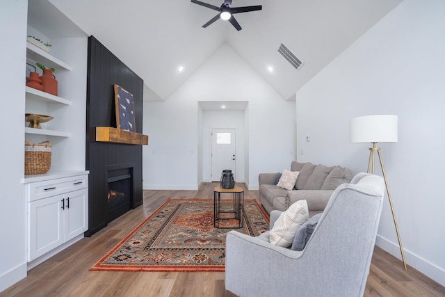 living room featuring built in shelves, wood-type flooring, high vaulted ceiling, a large fireplace, and ceiling fan