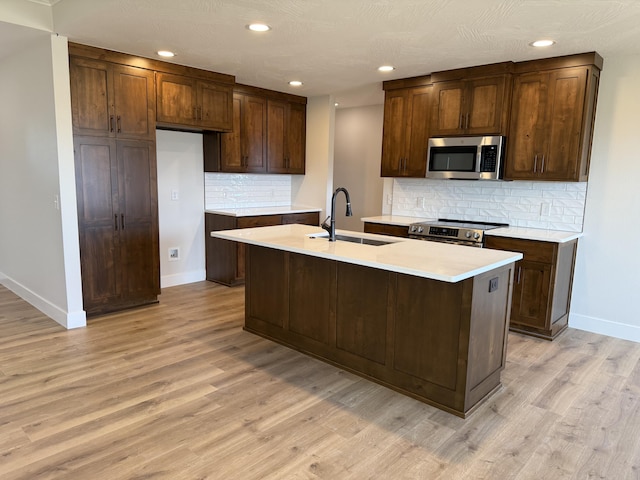 kitchen featuring sink, light wood-type flooring, appliances with stainless steel finishes, a kitchen island with sink, and decorative backsplash