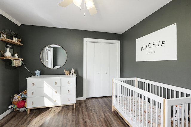 bedroom with a crib, dark wood-type flooring, ceiling fan, and a closet