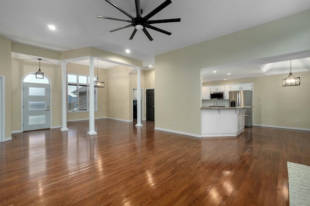 unfurnished living room with ornate columns, sink, ceiling fan with notable chandelier, and dark hardwood / wood-style flooring