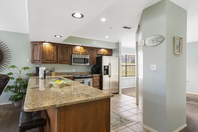 kitchen with light tile patterned floors, appliances with stainless steel finishes, light stone counters, a kitchen bar, and kitchen peninsula