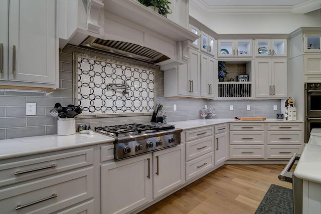 kitchen with appliances with stainless steel finishes, ornamental molding, white cabinets, custom exhaust hood, and light wood-type flooring