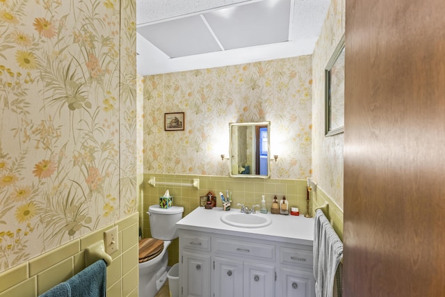 bathroom with toilet, a wainscoted wall, tile walls, vanity, and wallpapered walls