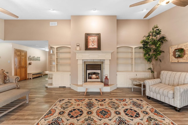 living area featuring built in features, visible vents, a ceiling fan, a glass covered fireplace, and wood finished floors