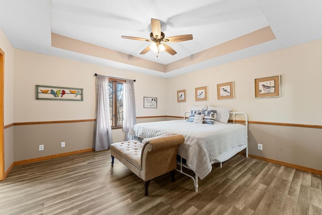 bedroom featuring a raised ceiling, ceiling fan, baseboards, and wood finished floors