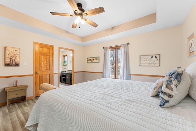 bedroom featuring ceiling fan, ensuite bath, light hardwood / wood-style floors, and a tray ceiling