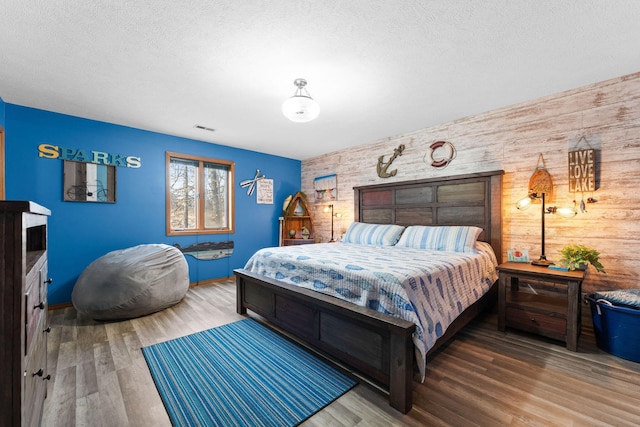 bedroom featuring wooden walls, visible vents, an accent wall, wood finished floors, and a textured ceiling