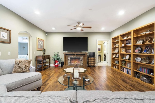 living room with arched walkways, a stone fireplace, a ceiling fan, and hardwood / wood-style floors