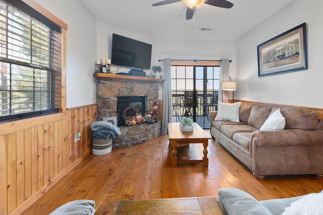 living room featuring wood-type flooring, a stone fireplace, ceiling fan, and wood walls