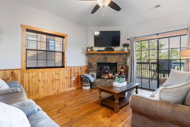 living room with a stone fireplace, hardwood / wood-style floors, ceiling fan, and wood walls