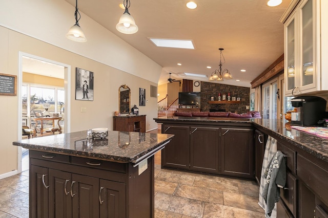 kitchen with a kitchen island, vaulted ceiling with skylight, pendant lighting, dark stone counters, and dark brown cabinets