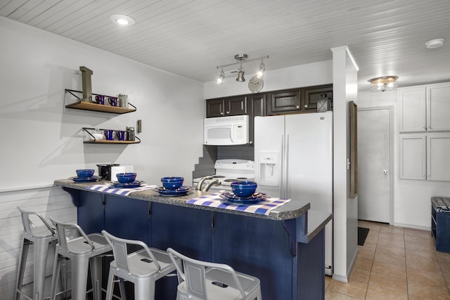 kitchen featuring a kitchen bar, dark brown cabinetry, light tile patterned floors, kitchen peninsula, and white appliances