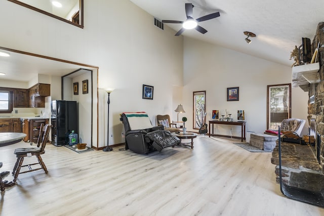 living room with ceiling fan, a stone fireplace, high vaulted ceiling, and light hardwood / wood-style flooring