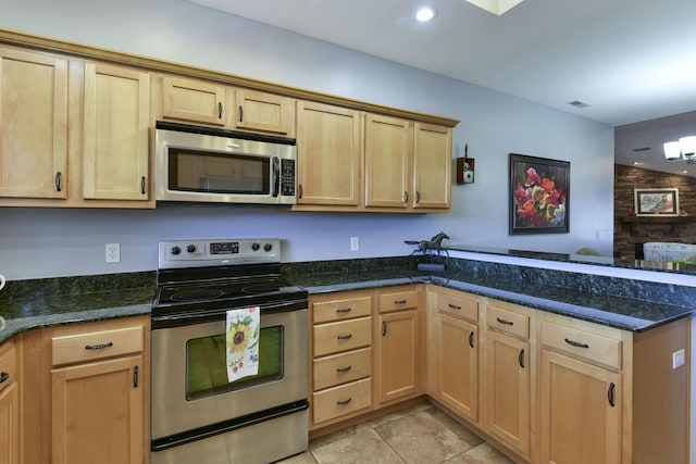 kitchen with appliances with stainless steel finishes, kitchen peninsula, light tile patterned flooring, light brown cabinetry, and dark stone counters