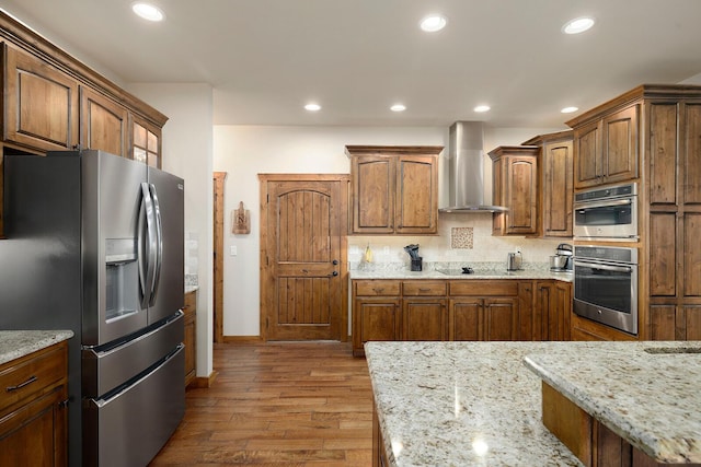 kitchen with backsplash, stainless steel appliances, light stone countertops, wall chimney exhaust hood, and light wood-type flooring