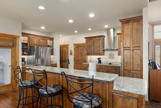 kitchen with a large island, dark wood-type flooring, stainless steel fridge, light stone countertops, and wall chimney exhaust hood