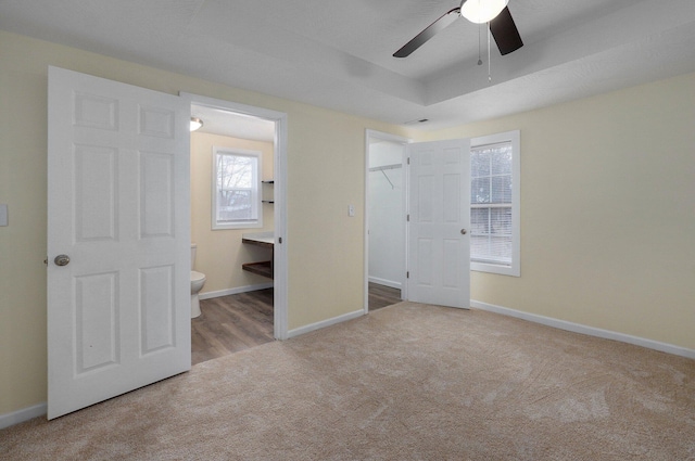 unfurnished bedroom with ceiling fan, light colored carpet, a tray ceiling, and ensuite bath