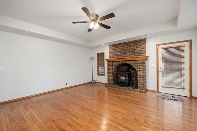 unfurnished living room with a raised ceiling, ceiling fan, a wood stove, and light wood-type flooring