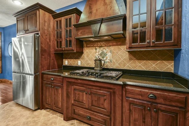 kitchen with light tile patterned floors, appliances with stainless steel finishes, tasteful backsplash, custom range hood, and dark stone counters