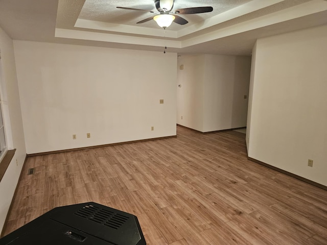 empty room with a tray ceiling, light wood finished floors, a ceiling fan, a textured ceiling, and baseboards