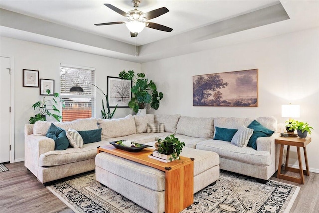 living room with a tray ceiling, baseboards, light wood-style floors, and a ceiling fan