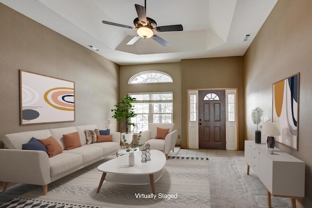 living area featuring visible vents, a ceiling fan, a textured wall, a tray ceiling, and light tile patterned flooring