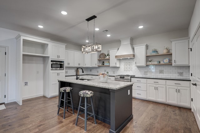 kitchen with an island with sink, appliances with stainless steel finishes, custom range hood, dark stone countertops, and white cabinets