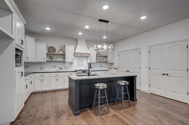 kitchen featuring a kitchen island with sink, sink, custom range hood, white cabinets, and pendant lighting