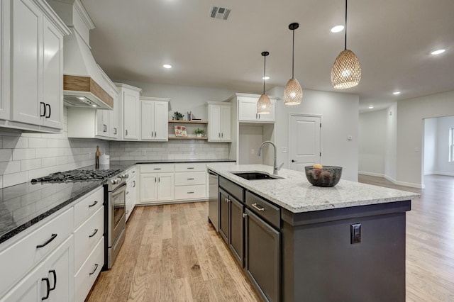 kitchen featuring appliances with stainless steel finishes, sink, premium range hood, hanging light fixtures, and a kitchen island with sink