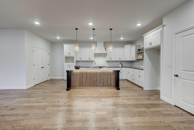 kitchen with light hardwood / wood-style flooring, pendant lighting, custom range hood, white cabinets, and an island with sink