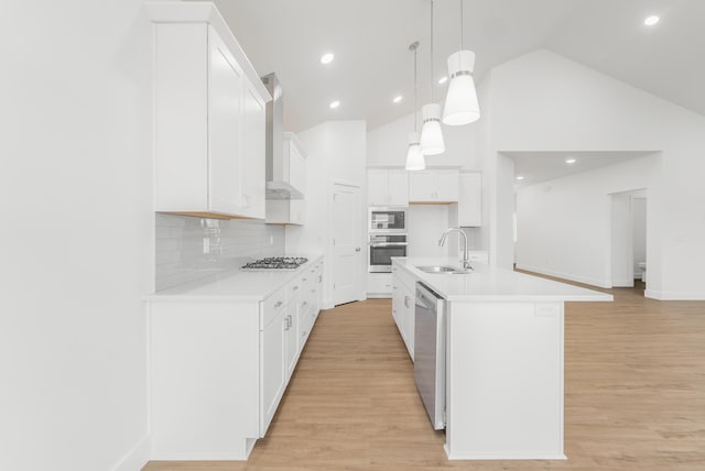 kitchen featuring decorative light fixtures, wall chimney range hood, stainless steel appliances, white cabinets, and a kitchen island with sink