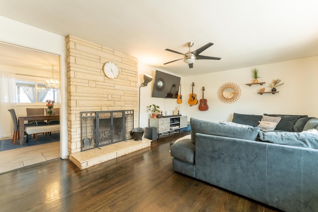 living area with ceiling fan with notable chandelier, a stone fireplace, and dark wood finished floors