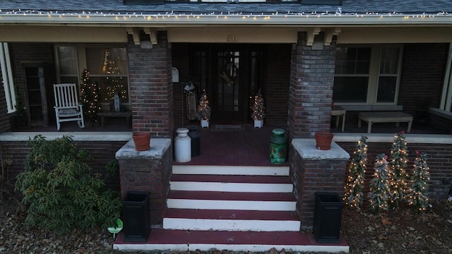 view of exterior entry featuring a porch, brick siding, and roof with shingles