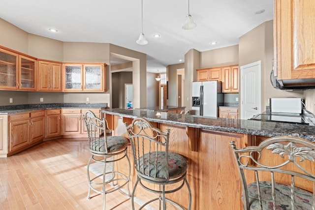 kitchen featuring stainless steel fridge, dark stone counters, glass insert cabinets, a kitchen breakfast bar, and pendant lighting