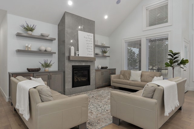 living room with high vaulted ceiling, a tiled fireplace, light wood-style flooring, and baseboards